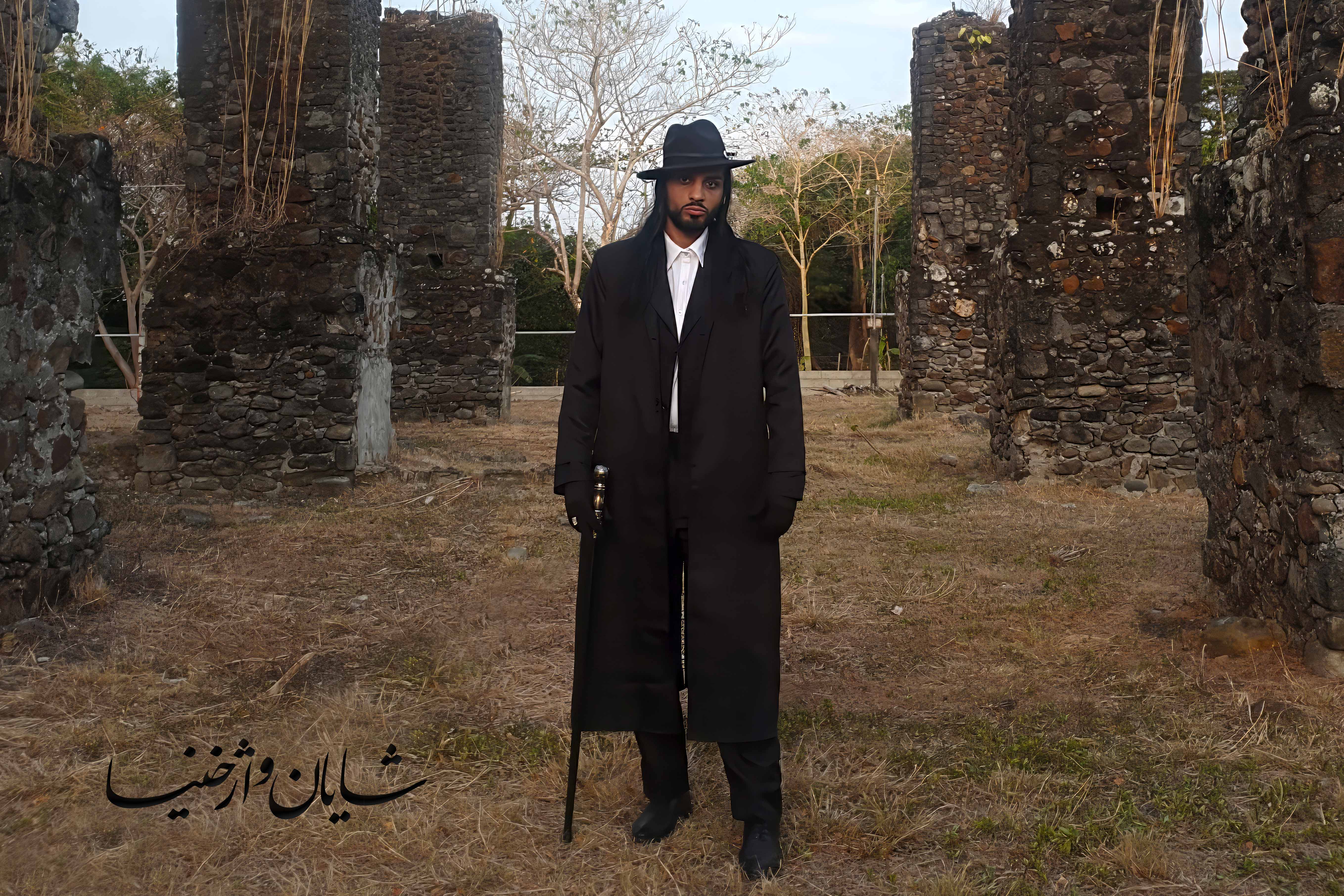 شایان در دوران معاصر با لباس مشکی ایستاده در کنار ویرانه‌های تاریخی